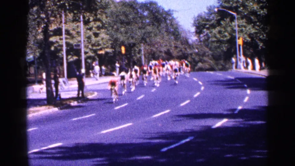The cycling group continues through the curve on a bright New York City street, with trees, signals, and roadside viewers visible.