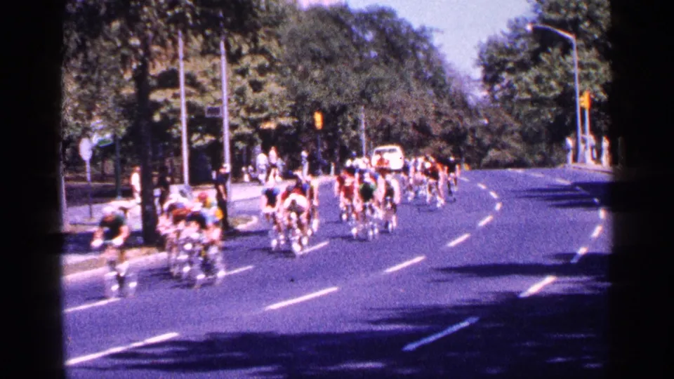 The cycling pack fills the curved New York City street as a white vehicle appears in the distance and spectators remain near the sidewalk.