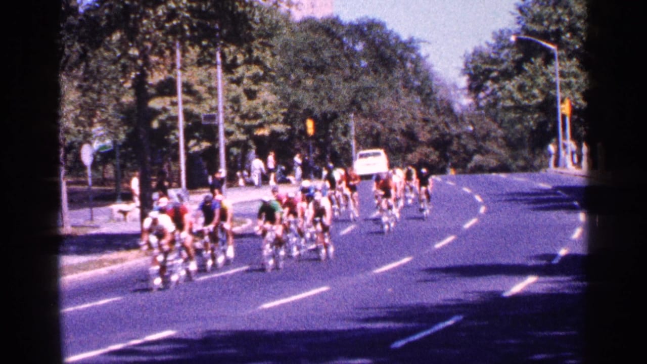 Cyclists ride in a pack along a curved park road with spectators nearby.