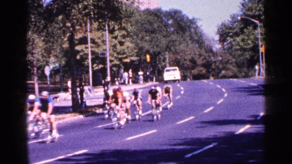 Cyclists in 1964 New York City ride away on a broad park road, with spectators and a white van visible near the tree-lined curb.
