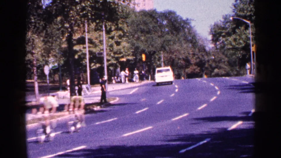 A trailing group of cyclists passes through the New York City park road scene, with trees, traffic lights, spectators, and a white van in view.
