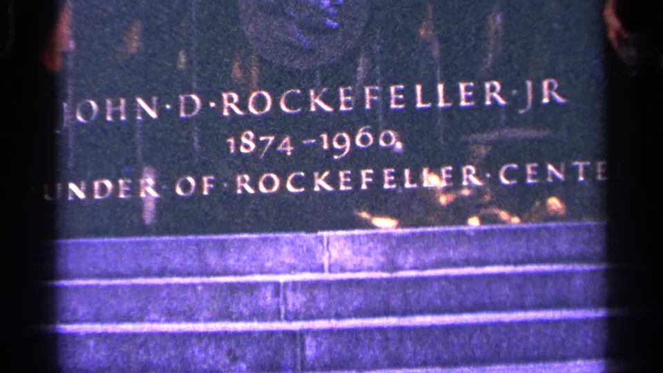 The John D. Rockefeller Jr. plaque remains in view, with bright water or reflections suggesting a nearby fountain setting.