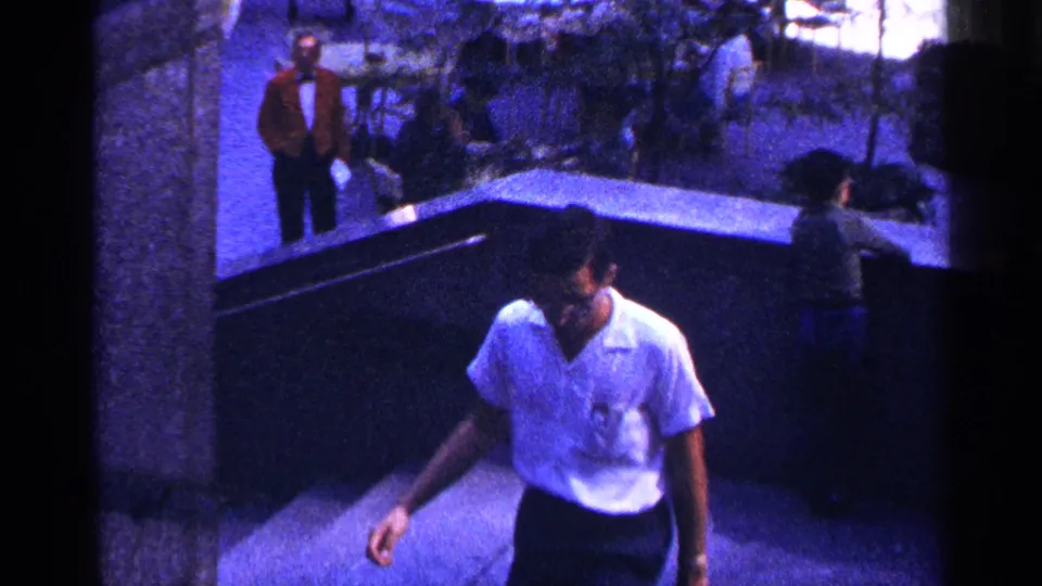 In 1964 New York City, a man in a white short-sleeve shirt walks up concrete stairs while pedestrians pass in the background.