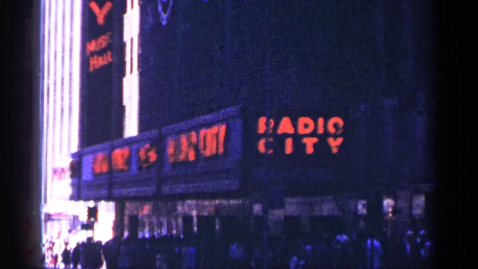 The red Radio City Music Hall signage appears on a dark New York City building facade with pedestrians below.