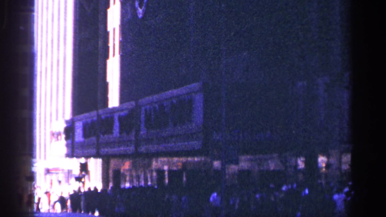 1964 New York City 8mm frame showing the Radio City Music Hall frontage at night with glowing marquee lights and people on the sidewalk.