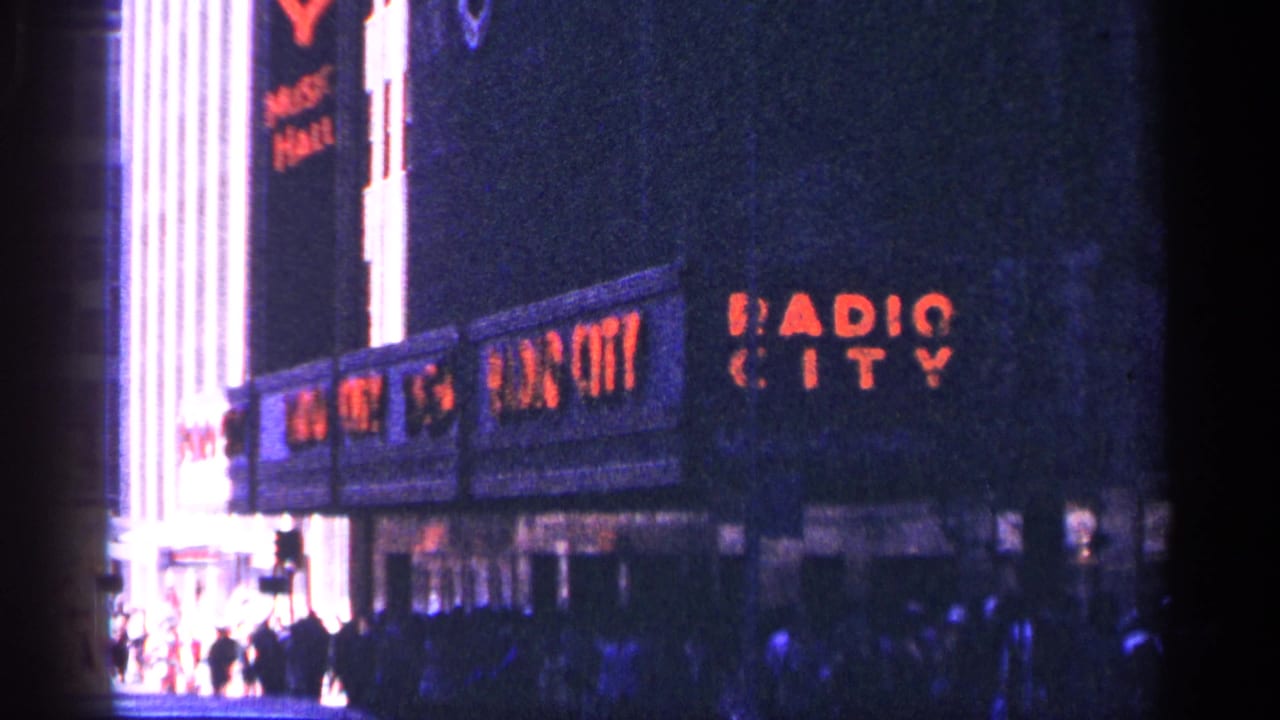1964 New York City 8mm frame with Radio City Music Hall lettering visible, bright marquee illumination, and a public crowd near the entrance.