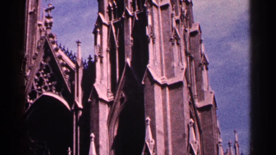 1964 New York City 8mm frame of a Gothic cathedral tower with pointed spires, carved stone panels, and blue sky.