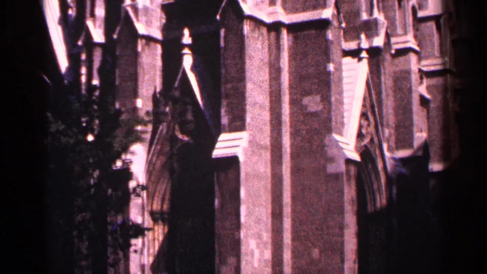 1964 New York City 8mm frame of a Gothic church wall with carved vertical bays, pointed arch detail, and dark foliage along the lower edge.