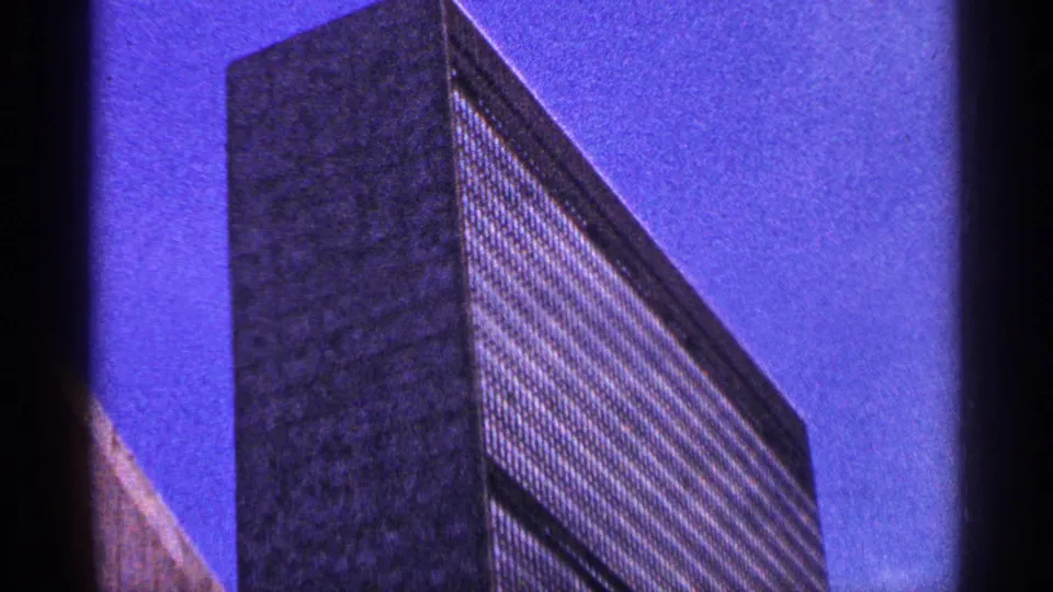 Low-angle New York City home-movie view of a modern skyscraper facade, framed by blue sky and 8mm gate edges.