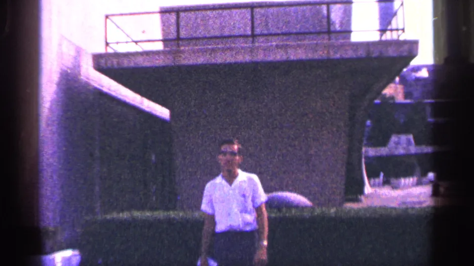 Adult man stands near hedges beneath a large concrete platform or wall in 1964 New York City home-movie footage.