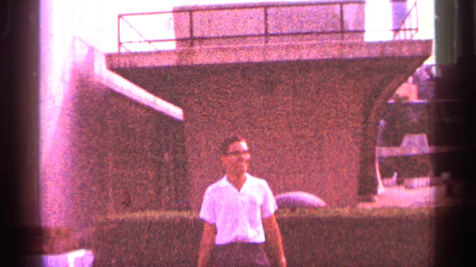 Red color cast over a 1964 New York City frame of an adult man posing near hedges and concrete architecture.
