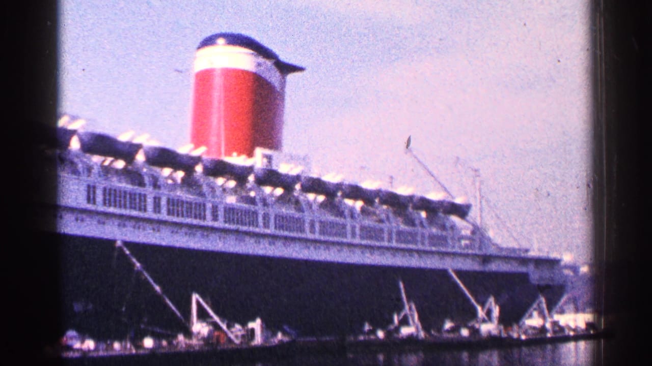 Large ocean liner docked beside harbor water with red funnel and white railings visible.