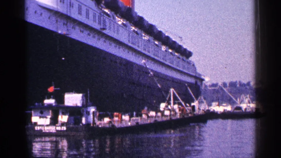 From a closer dockside angle, the liner's dark hull towers above tugboats, cranes, and calm harbor water in 1964 New York City.