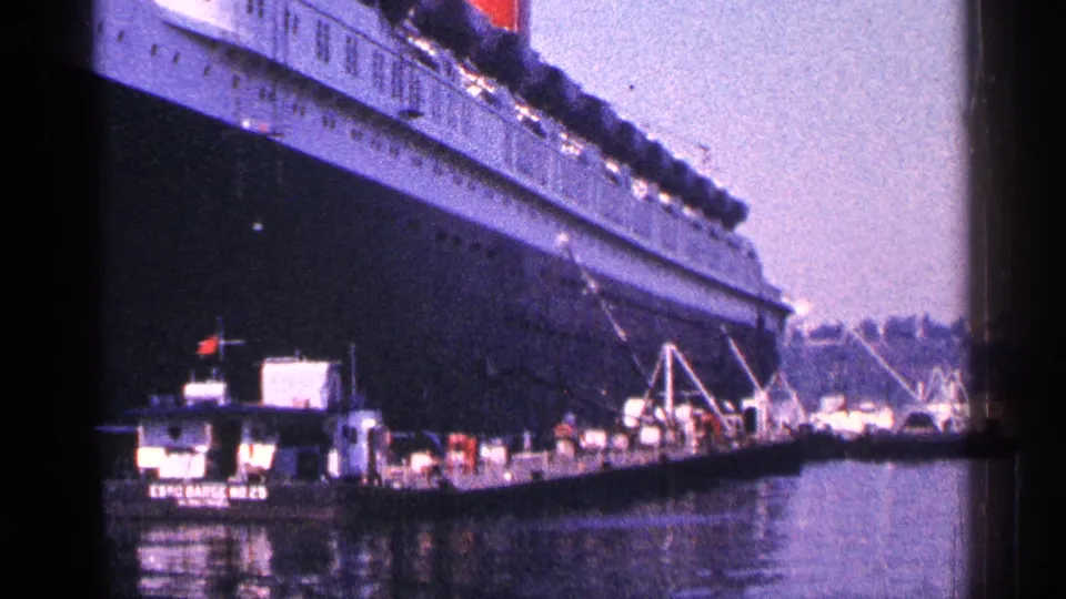 A dockside 1964 New York City harbor frame shows the ocean liner's hull above tugboats, pier machinery, and reflective water.