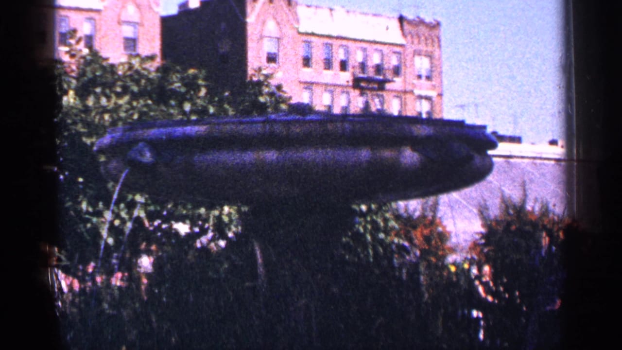 Dark saucer-shaped fountain with water spray, shrubs, and brick apartment buildings behind it.