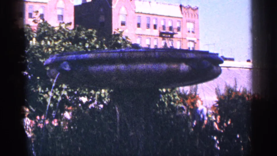 A static 1964 home-movie frame appears to show a New York City park fountain or monument set above shrubs, with distant visitors and brick buildings behind.