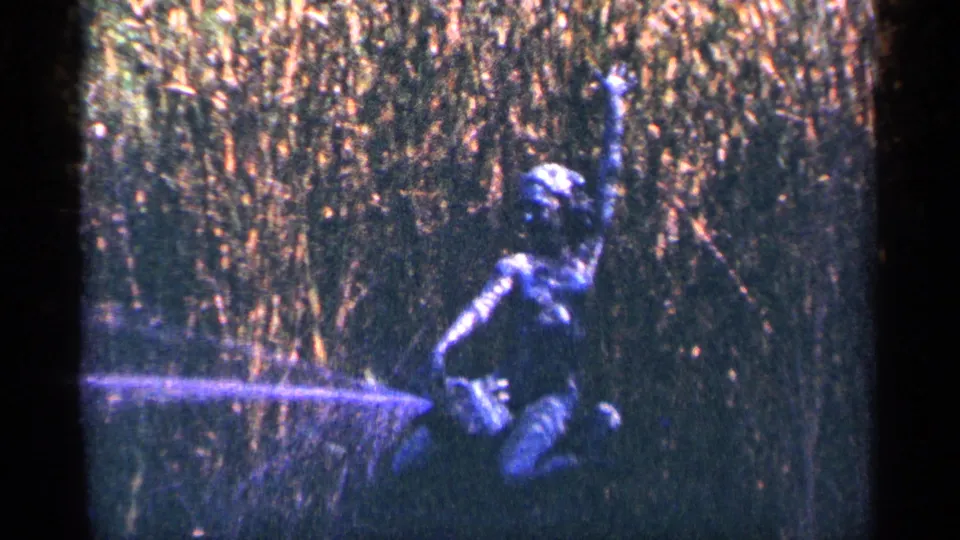 A dark kneeling garden statue or figure appears near a low water spray, backed by tall reeds or dense grasses.