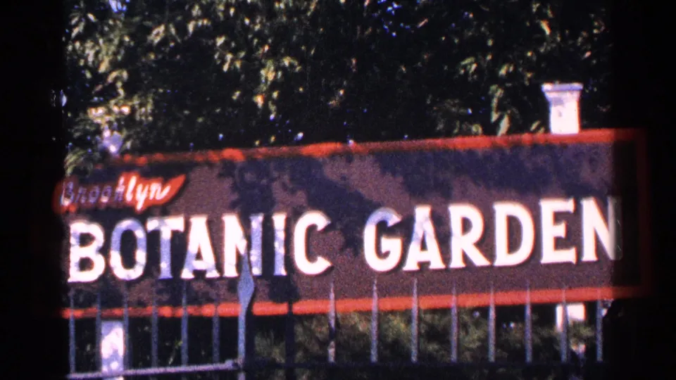 1964 New York City frame of the Brooklyn Botanic Garden sign, with bold white lettering, red trim, fence rails, and foliage.