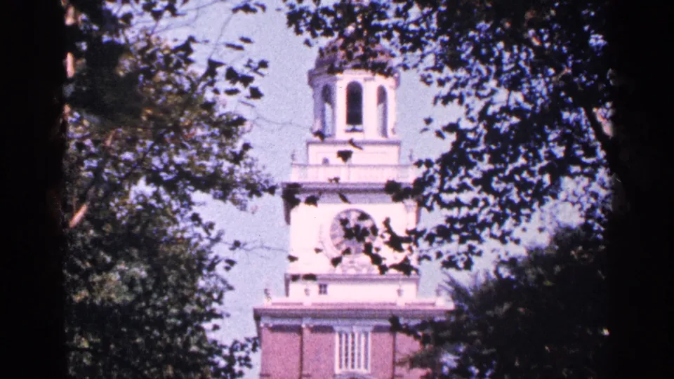 1964 New York City home-movie frame showing a pale clock tower and brick building partly obscured by tree branches.