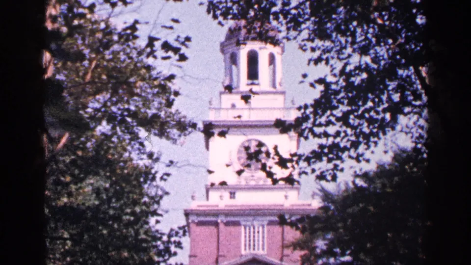 1964 New York City frame of a clock tower above a brick facade, partly hidden by dark leaves and branches.