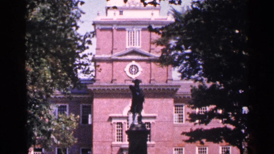 1964 8mm view of a historic brick landmark, with a dark statue on a pedestal and leafy trees partly obscuring the facade.