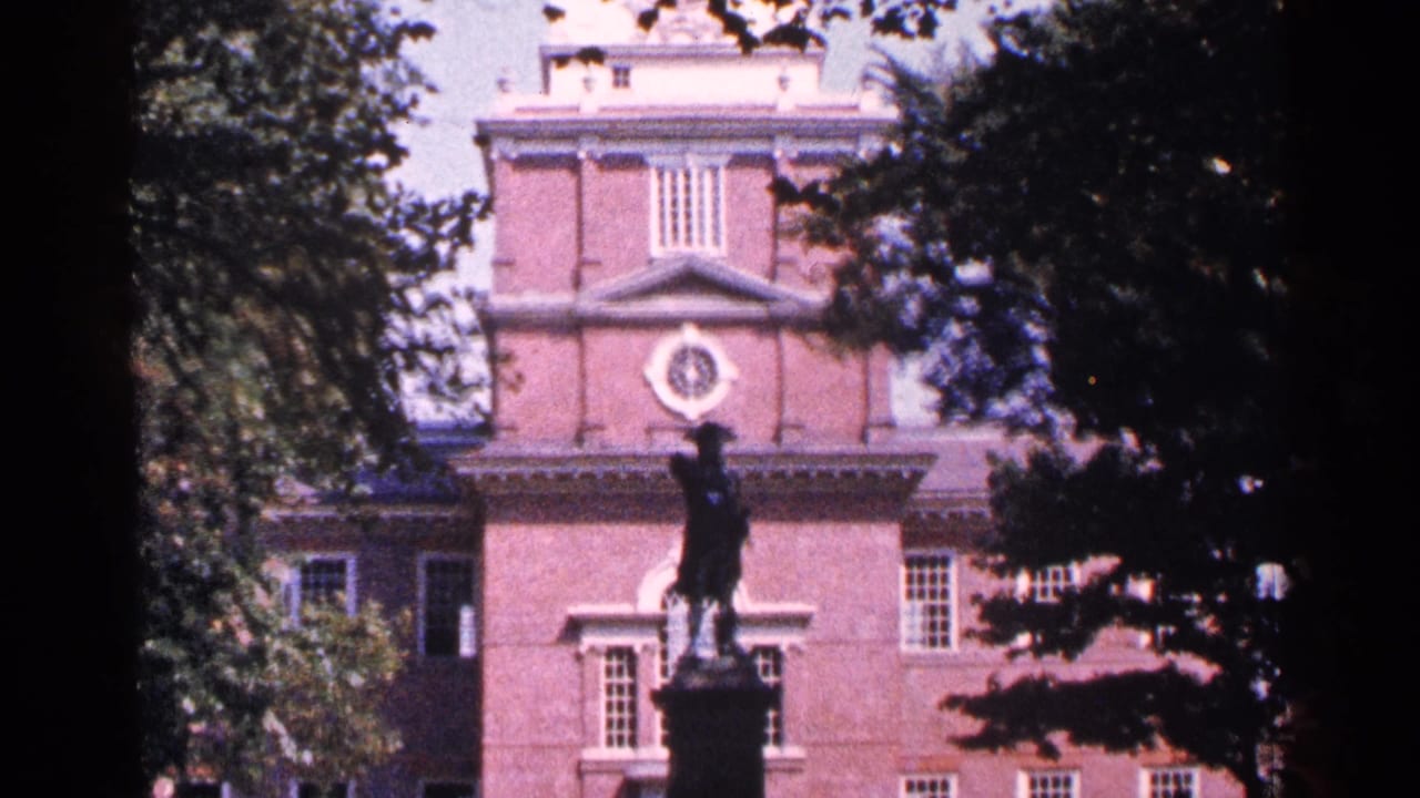 Historic brick facade behind a statue, framed by tree branches and shade.