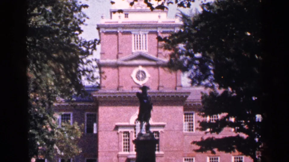 1964 home-movie frame showing a red brick historic building, statue silhouette, white architectural trim, and trees around the view.