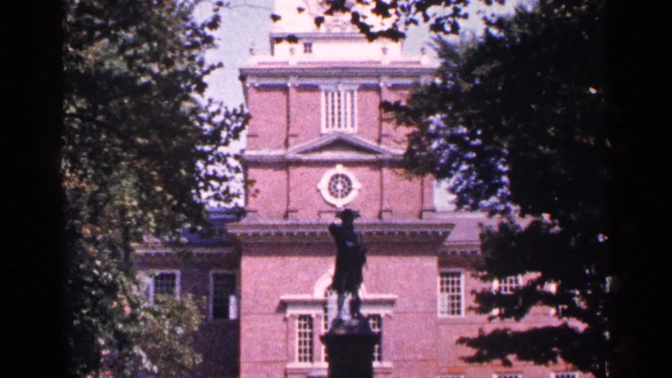 1964 8mm frame with a centered statue in front of a historic brick building, softened by film grain and tree cover.