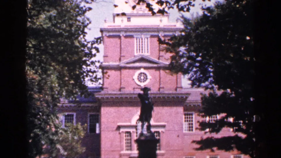 1964 archival home-movie still of a historic brick landmark with a foreground statue, white trim, and dense trees along the frame edges.