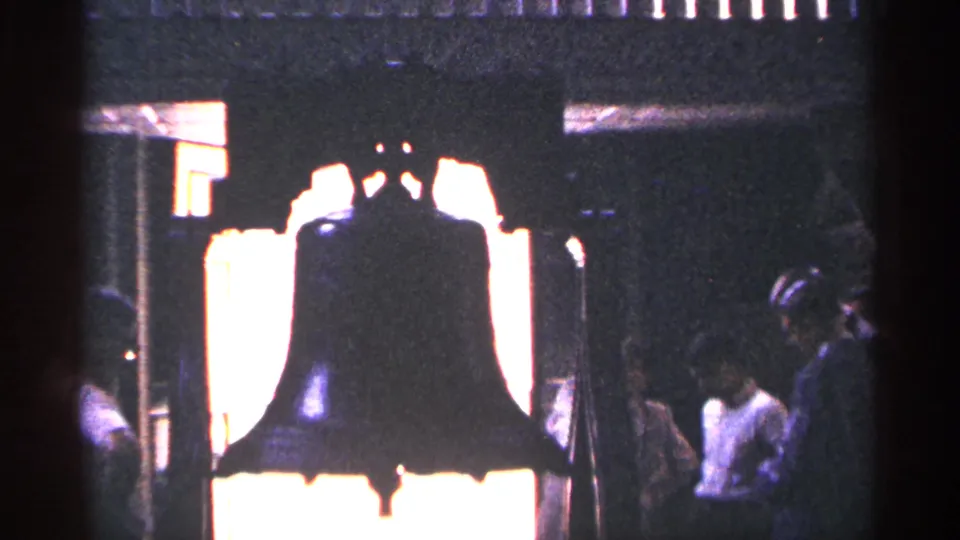 The Liberty Bell appears in silhouette against bright light, with visitors visible to the side inside the historic display area.