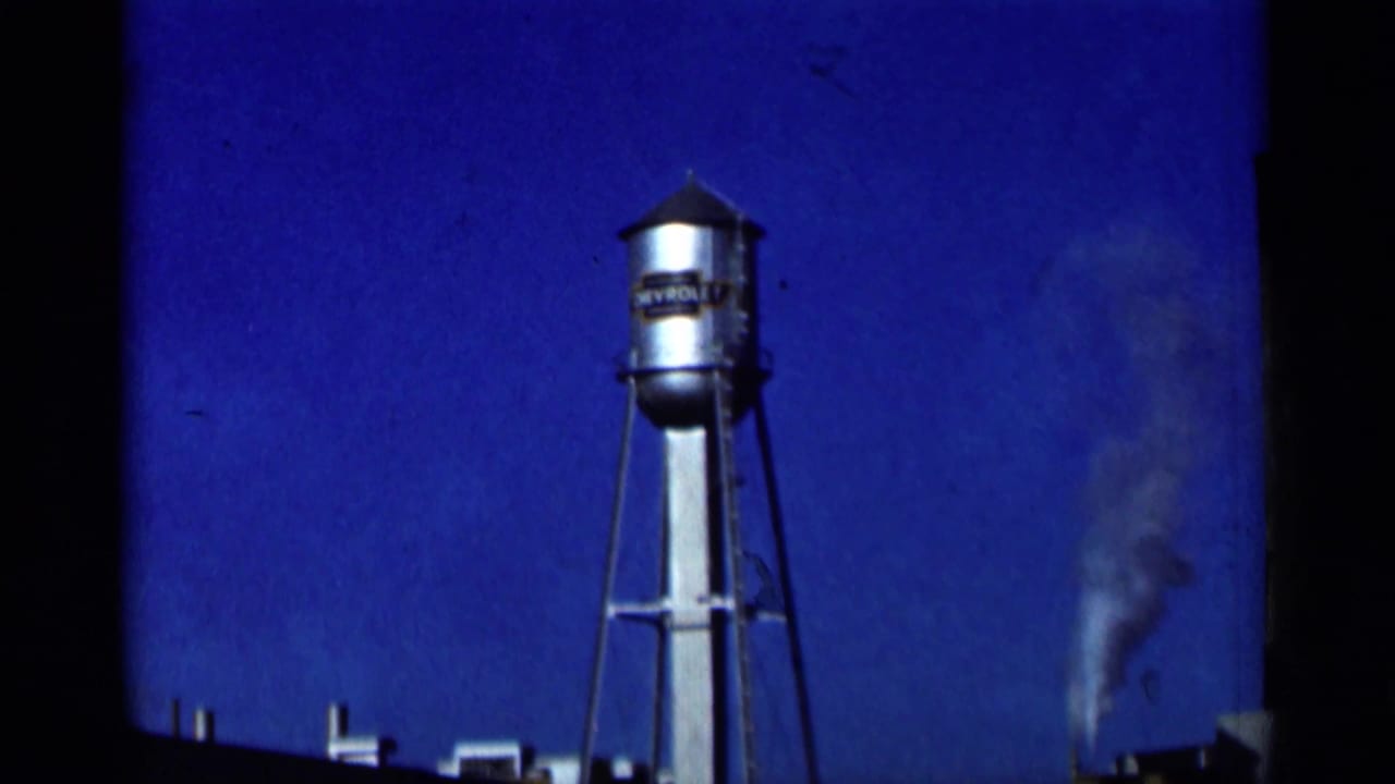 1964 8mm frame showing a Chevrolet-marked water tower, blue sky, film edge, and smoke rising in the distance.