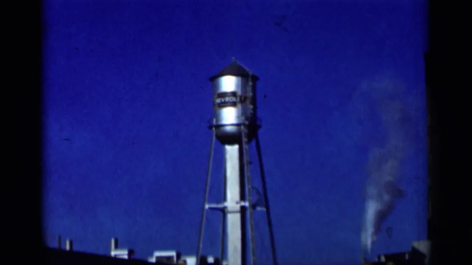 1964 8mm frame showing a Chevrolet-marked water tower, blue sky, film edge, and smoke rising in the distance.