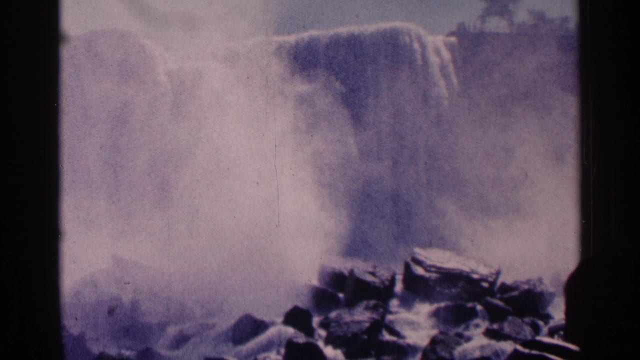 1964 8mm view of Niagara Falls, Canada, with mist rising behind dark rocks and visible film-frame borders.