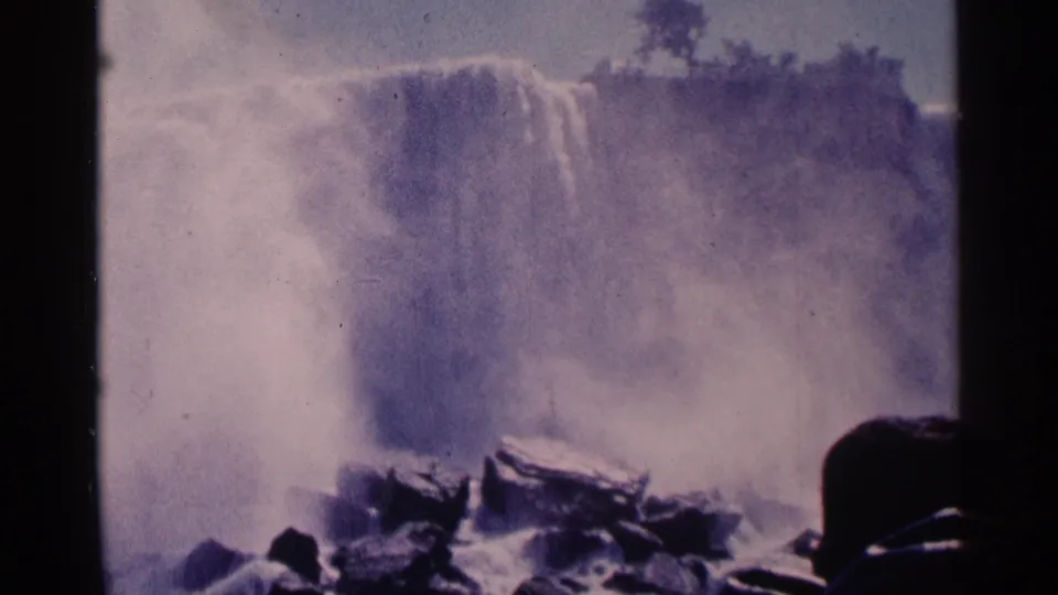 1964 8mm frame of Niagara Falls, Canada, with a hooded visitor silhouetted against mist and falling water.