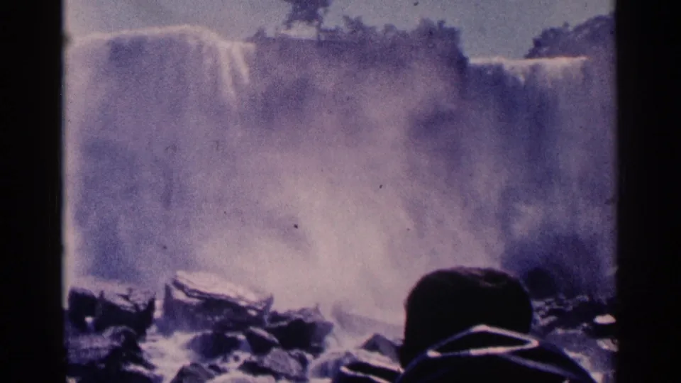 1964 8mm Niagara Falls view with dense spray, dark foreground rocks, and a hooded visitor partly visible below.