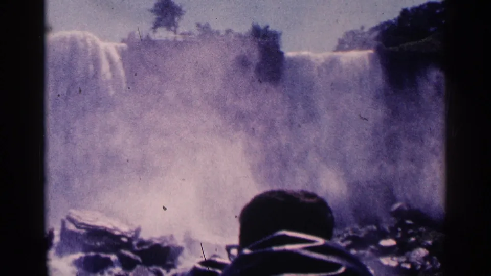 1964 8mm frame of Niagara Falls, Canada, showing misty cascades, dark rocks, and a hooded observer from behind.