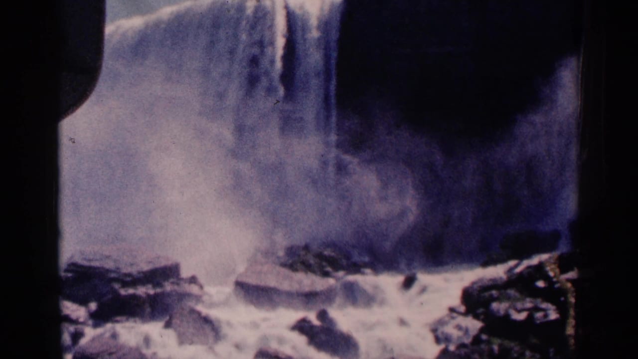 1964 8mm view of Niagara Falls, Canada, with water dropping over a cliff, spray rising, and rocks in the foreground.