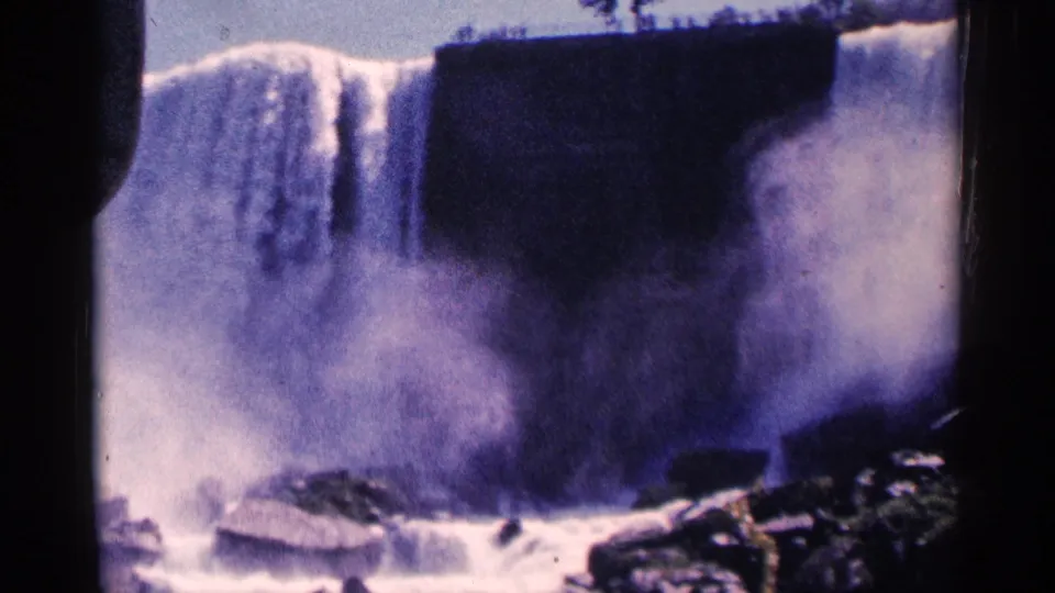 1964 Niagara Falls travel-film frame showing the falls, rising spray, rocks below, and small visitors along the upper rim.