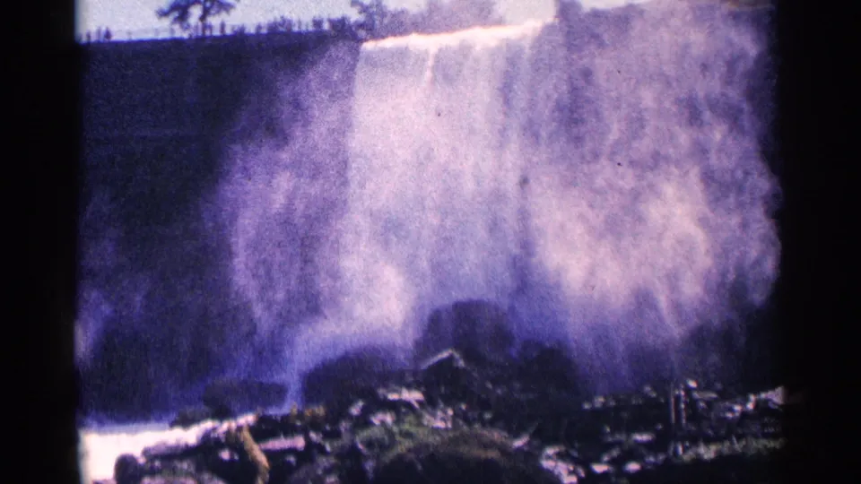 1964 Niagara Falls, Canada, home-movie frame with a broad curtain of mist, rocks at the base, and tiny visitors on the rim.