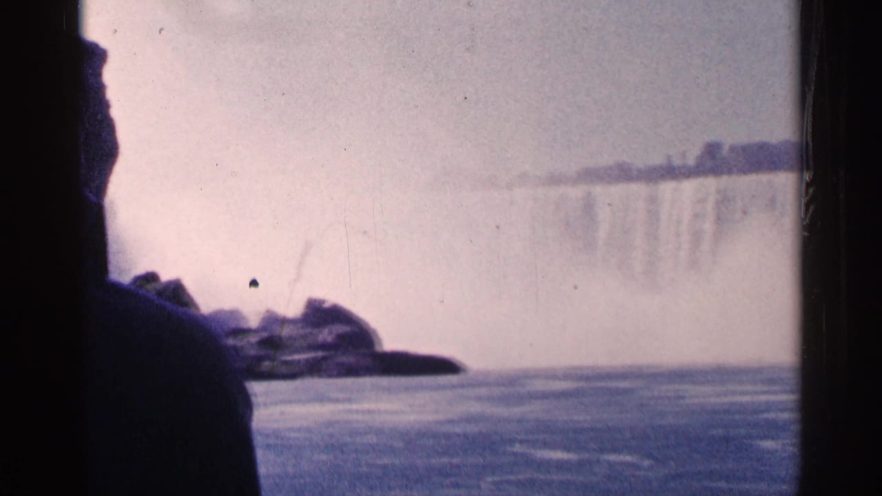 1964 8mm view of Niagara Falls with a dark foreground figure, rocky edge, river surface, mist, and waterfall in hazy daylight.