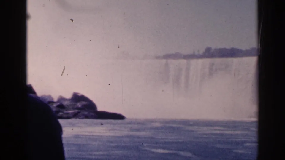 Niagara Falls appears beyond dark structural framing, with a rocky foreground, smooth river water, mist, and pale sky.