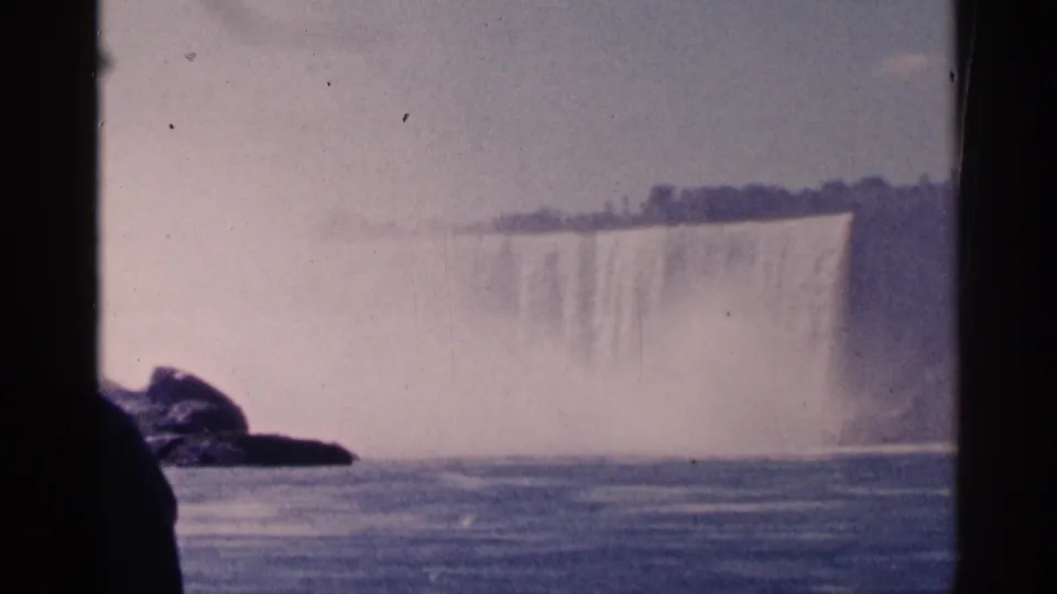 1964 home-movie frame shows Niagara Falls across the river, with mist rising behind rocks and dark shaded foreground edges.