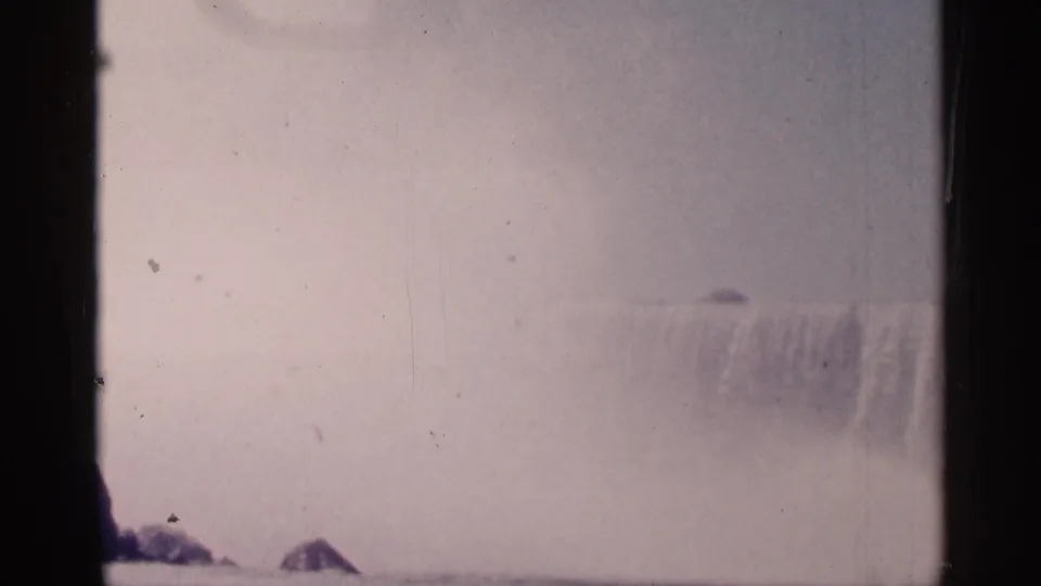 Wide 8mm frame of Niagara Falls, Canada, showing misty cascade, low rocks, faded color, and visible film wear.