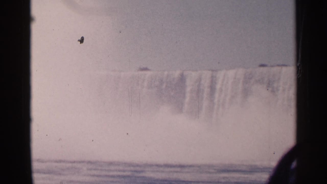 Misty waterfall crest with pale sky, river surface, film border, and small debris specks.