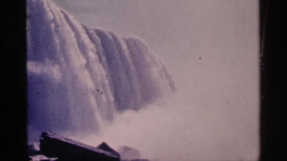 1964 Niagara Falls home-movie frame with the waterfall edge, rising mist, dark rocks, and faded purplish 8mm color.