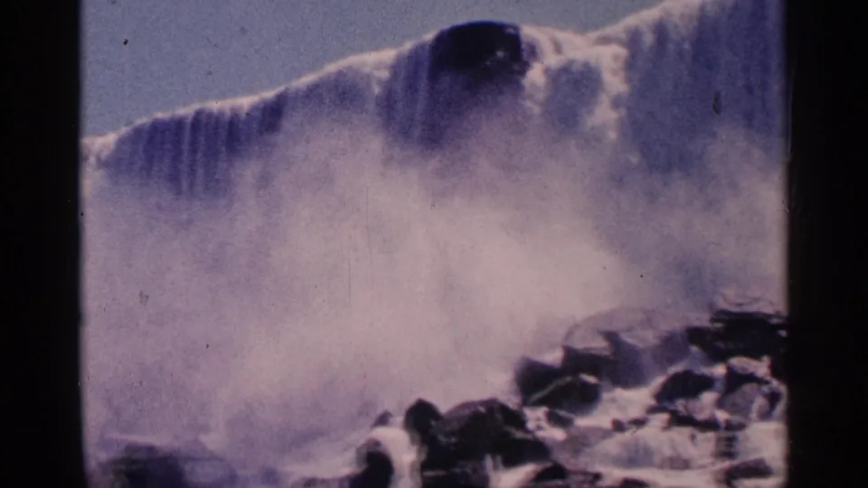 1964 8mm frame of Niagara Falls with the cascade softened by mist and dark rocks visible along the lower right.