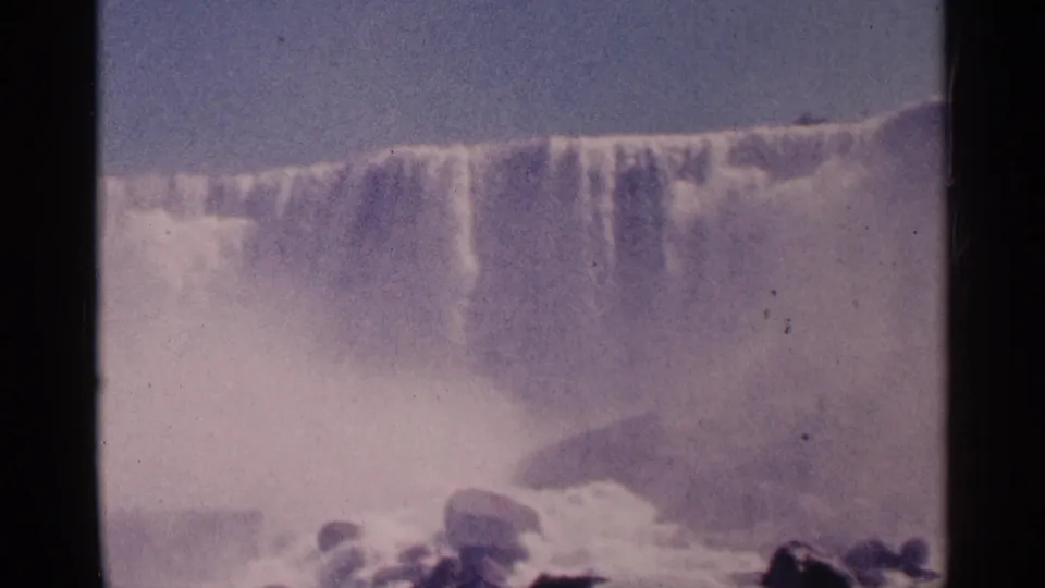 1964 8mm view of Niagara Falls with a distant waterfall curtain, rising mist, and low foreground rocks.