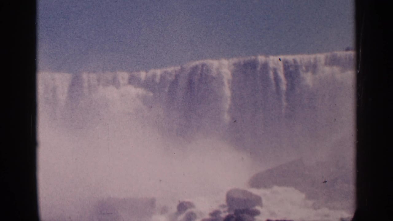 1964 8mm home-movie frame of Niagara Falls in Canada, with a wide waterfall crest, rocks, foam, and thick mist below.
