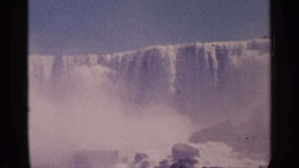1964 8mm home-movie frame of Niagara Falls in Canada, with a wide waterfall crest, rocks, foam, and thick mist below.