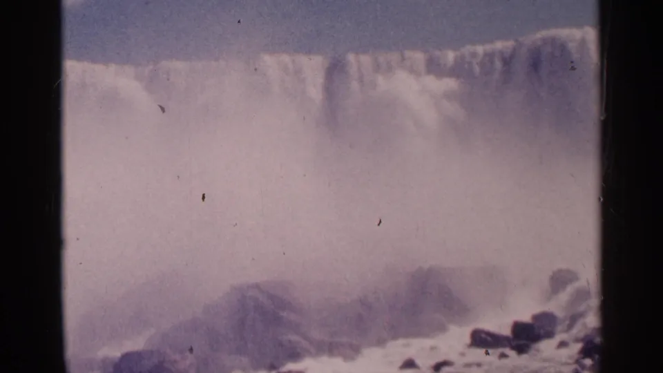 Vintage 8mm frame of Niagara Falls with the cascade softened by thick mist, pale water, rocks, and visible film grain.